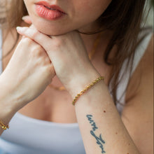 Load image into Gallery viewer, Woman with a tattoo on her wrist wearing a gold bracelet, sitting indoors.
